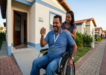 Homem em cadeira de rodas ergue chaves de imóvel sorrindo, ao lado de mulher que o abraça em frente à casa nova