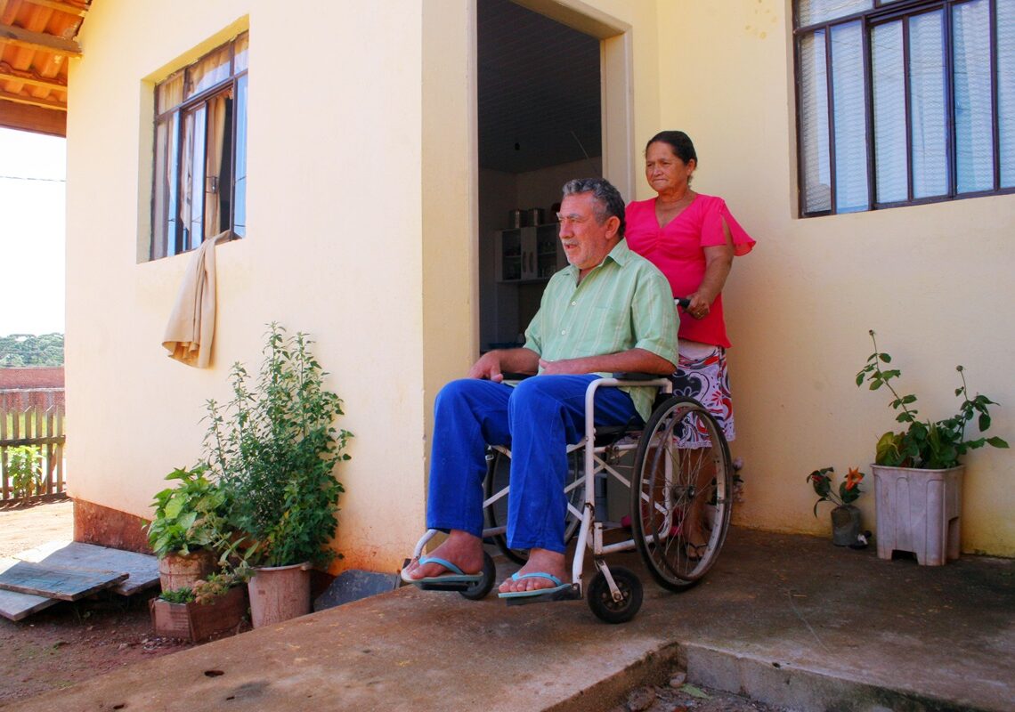Homem idoso em cadeira de rodas usa camisa verde e é acompanhado por mulher de blusa rosa na varanda de casa simples