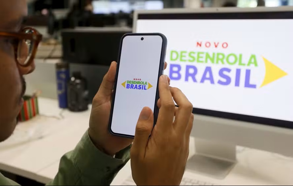Pessoa segurando celular com logo Novo Desenrola Brasil, monitor ao fundo exibe mesma identidade visual colorida