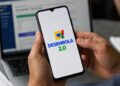 Mãos segurando smartphone com logo do Desenrola 2.0 na tela, notebook ao fundo em ambiente de escritório