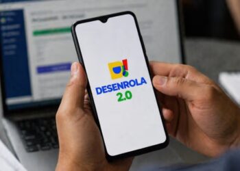 Mãos segurando smartphone com logo do Desenrola 2.0 na tela, notebook ao fundo em ambiente de escritório