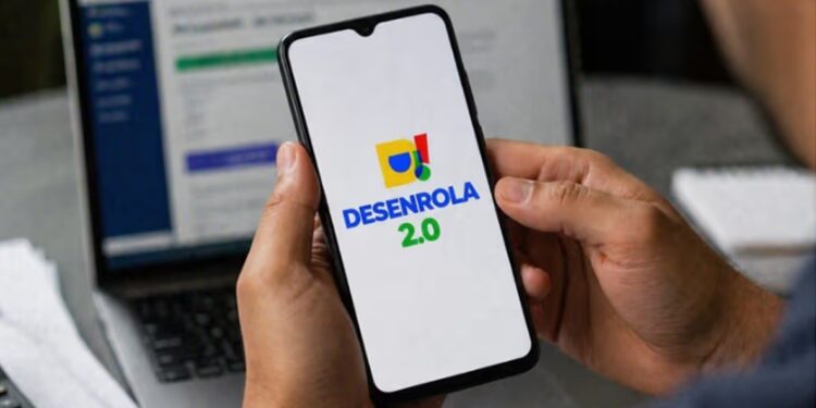Mãos segurando smartphone com logo do Desenrola 2.0 na tela, notebook ao fundo em ambiente de escritório