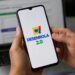 Mãos segurando smartphone com logo do Desenrola 2.0 na tela, notebook ao fundo em ambiente de escritório