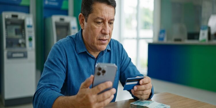 Homem de meia-idade usando camisa azul consulta celular e segura cartão em agência, com notas de R$ 100 na mesa