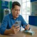 Homem de meia-idade usando camisa azul consulta celular e segura cartão em agência, com notas de R$ 100 na mesa