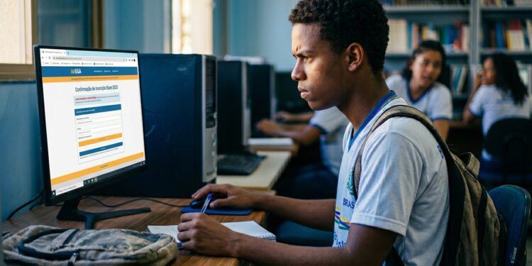 Jovem de uniforme escolar acessa sistema de confirmação do Enem 2026 em computador de laboratório com estantes de livros ao fundo