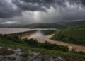 Barragem libera água barrenta sob nuvens escuras de tempestade, cercada por vegetação verde e plantações