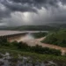 Barragem libera água barrenta sob nuvens escuras de tempestade, cercada por vegetação verde e plantações
