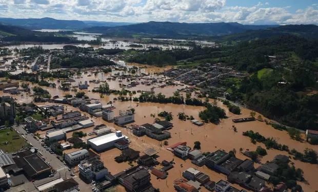 Vista aérea de cidade em Santa Catarina alagada por enchente, com água marrom cobrindo ruas e áreas urbanas cercadas de morros verdes