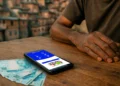 Mãos de homem apoiadas em mesa de madeira com celular mostrando app do Bolsa Família e notas de R$100