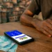 Mãos de homem apoiadas em mesa de madeira com celular mostrando app do Bolsa Família e notas de R$100