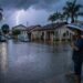 Homem com guarda-chuva listrado observa rua residencial alagada sob tempestade com raio ao fundo