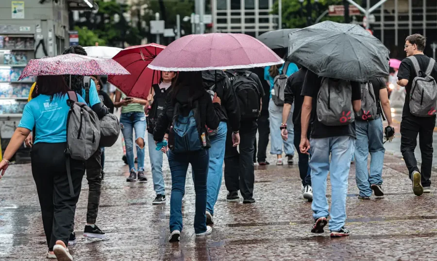 Pessoas caminham em calçamento molhado, segurando guarda-chuvas coloridos durante chuva intensa em área urbana