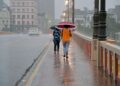 Duas pessoas caminham com guarda-chuvas vermelho e azul sobre ponte urbana molhada durante chuva intensa