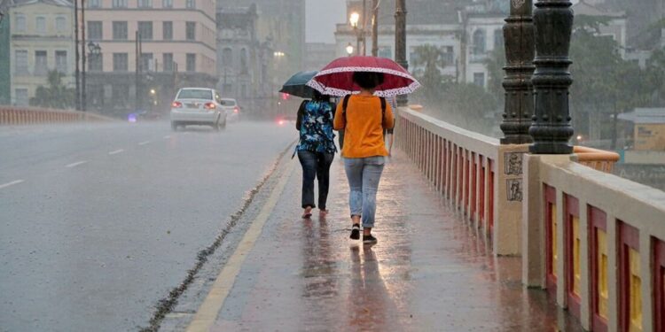 Duas pessoas caminham com guarda-chuvas vermelho e azul sobre ponte urbana molhada durante chuva intensa