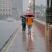Duas pessoas caminham com guarda-chuvas vermelho e azul sobre ponte urbana molhada durante chuva intensa