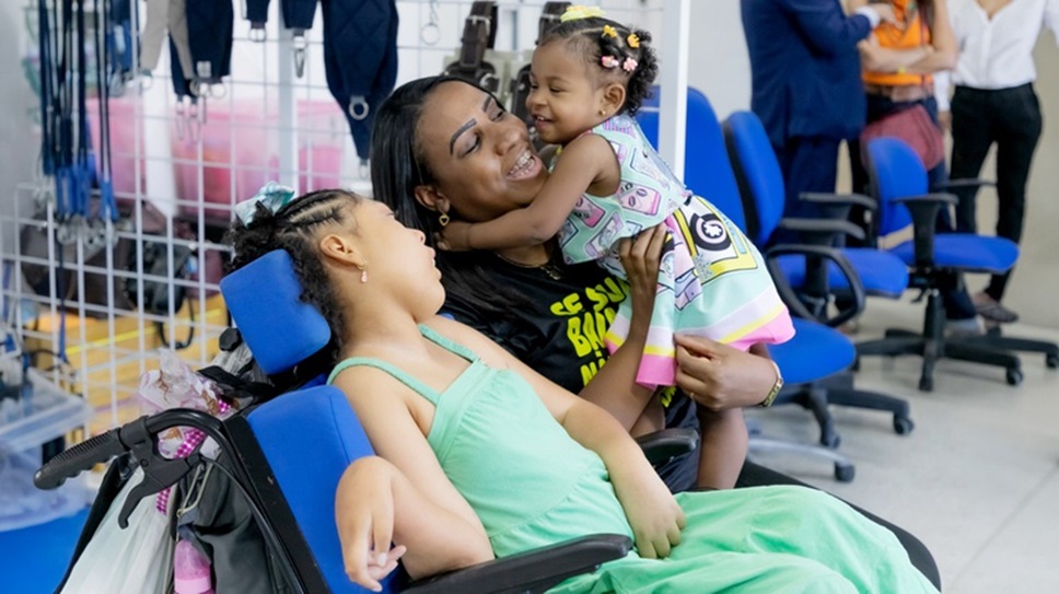 Mulher sorridente segura criança pequena enquanto outra criança com microcefalia está reclinada em cadeira de reabilitação