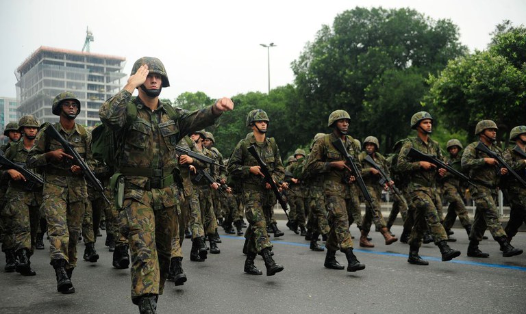 Soldados do Exército Brasileiro marchando armados com fuzis, usando uniformes camuflados e capacetes em via urbana
