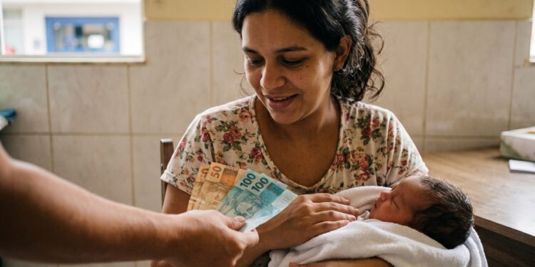 Mulher sorri segurando bebê recém-nascido enquanto recebe notas de reais de R$ 50 e R$ 100 em agência da CAIXA