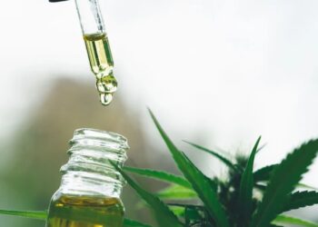 Conta-gotas com óleo dourado de CBD pingando em frasco de vidro, com folhas verdes de cannabis ao fundo em fundo desfocado