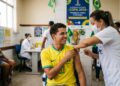 Jovem com camisa da Seleção Brasileira recebe vacina contra sarampo de profissional do SUS em posto de vacinação