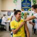 Jovem com camisa da Seleção Brasileira recebe vacina contra sarampo de profissional do SUS em posto de vacinação