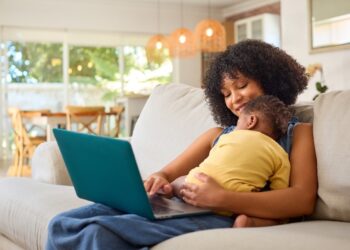 Mulher negra de cabelos cacheados usa notebook azul-petróleo com criança de blusa amarela no colo, sentada em sofá bege