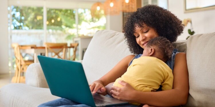Mulher negra de cabelos cacheados usa notebook azul-petróleo com criança de blusa amarela no colo, sentada em sofá bege