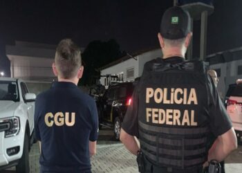 Agentes da CGU e Polícia Federal de costas durante operação noturna com viaturas ao fundo em área residencial