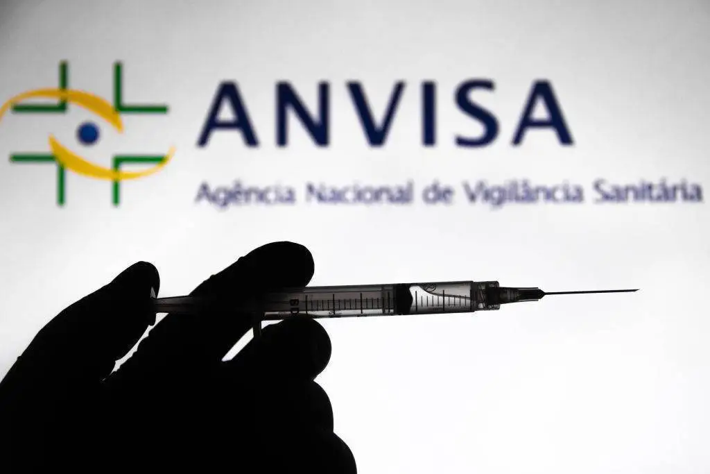 Mão segura seringa com líquido transparente em silhueta contra fundo iluminado com logotipo da Anvisa