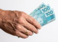 Mão idosa segura 300 reais em notas de R$100.