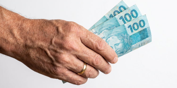 Mão idosa segura 300 reais em notas de R$100.