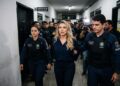 Deolane Bezerra, loira de macaquinho azul marinho, escoltada por dois policiais civis em corredor de delegacia
