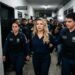 Deolane Bezerra, loira de macaquinho azul marinho, escoltada por dois policiais civis em corredor de delegacia