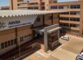 Fachada da sede da Anvisa com logo verde e amarelo, edifício bege com cobertura metálica e estacionamento