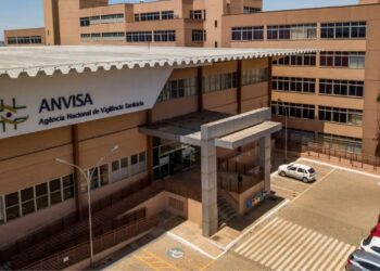 Fachada da sede da Anvisa com logo verde e amarelo, edifício bege com cobertura metálica e estacionamento