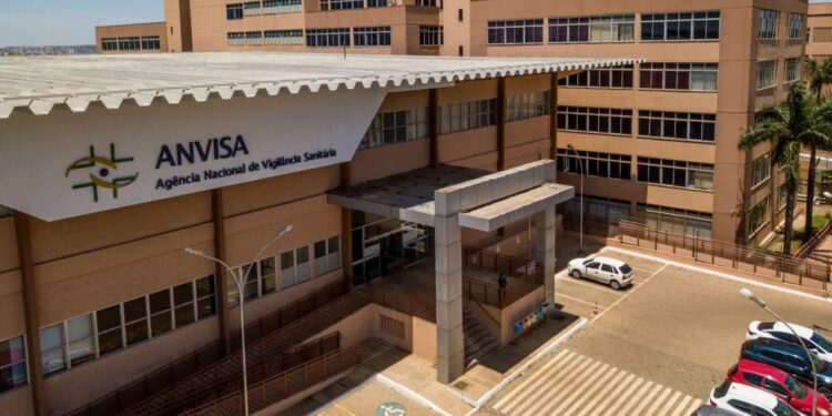 Fachada da sede da Anvisa com logo verde e amarelo, edifício bege com cobertura metálica e estacionamento