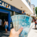 Notas de R$ 100 em frente à agência Caixa em rua comercial.