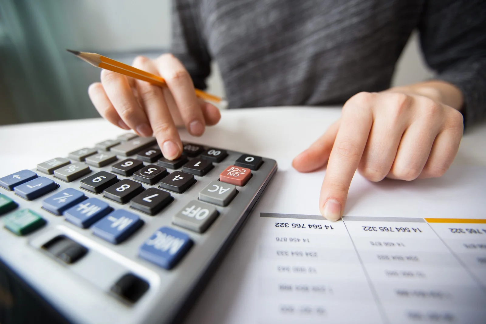 Mãos usando calculadora científica com lápis amarelo e planilha de dados numéricos sobre mesa branca
