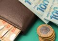 Carteira de couro marrom com notas de R$ 100 e R$ 50, além de moedas de R$ 1 empilhadas sobre fundo verde