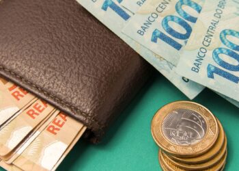 Carteira de couro marrom com notas de R$ 100 e R$ 50, além de moedas de R$ 1 empilhadas sobre fundo verde