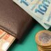 Carteira de couro marrom com notas de R$ 100 e R$ 50, além de moedas de R$ 1 empilhadas sobre fundo verde