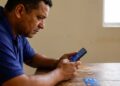 Homem de meia-idade com camisa polo azul consulta aplicativo financeiro no celular, cartão Caixa sobre mesa de madeira