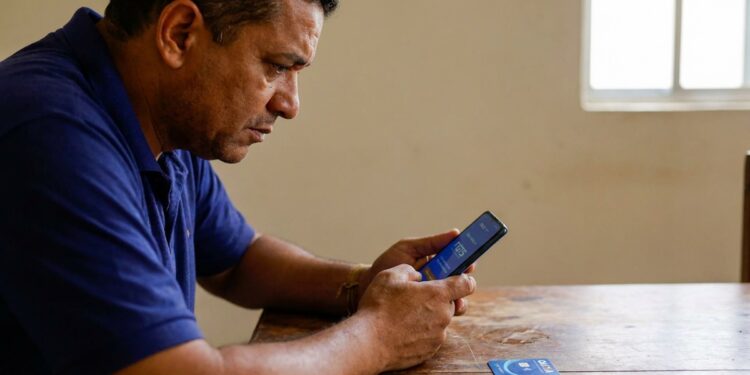 Homem de meia-idade com camisa polo azul consulta aplicativo financeiro no celular, cartão Caixa sobre mesa de madeira