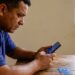 Homem de meia-idade com camisa polo azul consulta aplicativo financeiro no celular, cartão Caixa sobre mesa de madeira