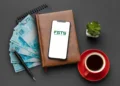 Notas de 100 reais, smartphone exibindo logo do FGTS sobre agenda de couro, caneta, xícara de café e suculenta