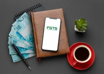 Notas de 100 reais, smartphone exibindo logo do FGTS sobre agenda de couro, caneta, xícara de café e suculenta
