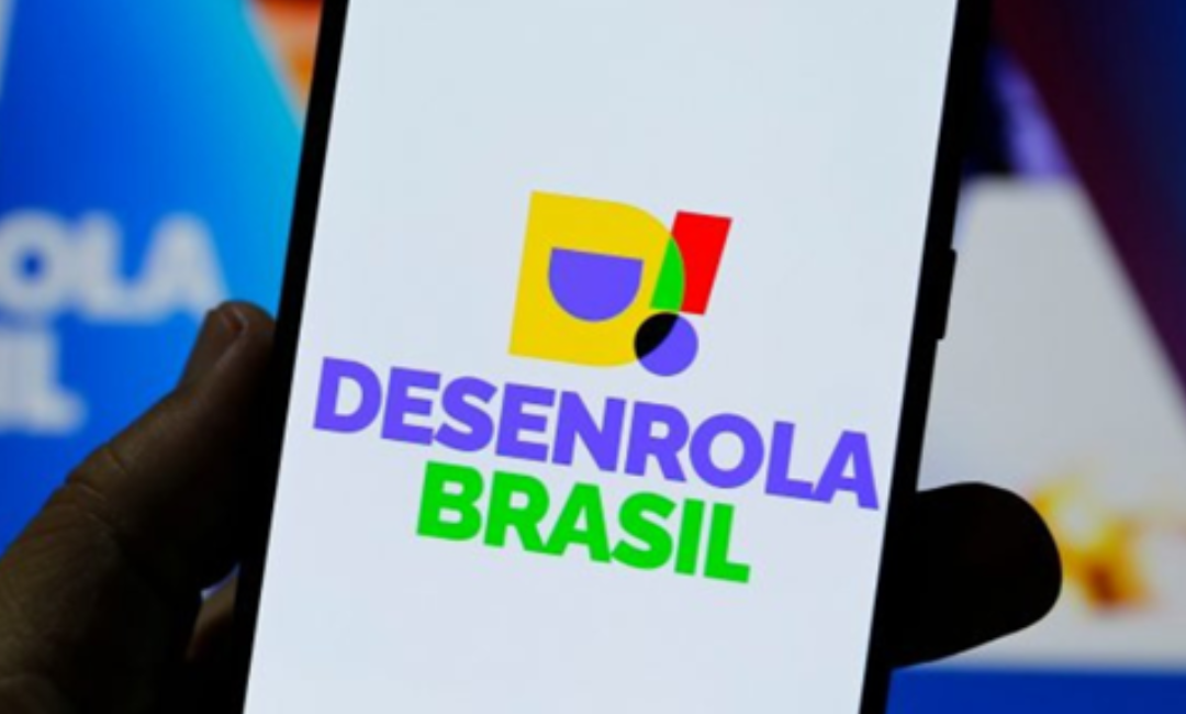 Mão segurando celular com logo colorido do programa Desenrola Brasil na tela em fundo azul desfocado