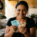 Mulher conta notas de real sorrindo dentro de casa e, ao redor, um sofá e a cozinha de uma residência simples