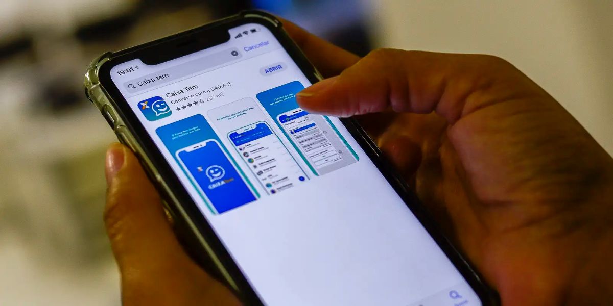 Mãos seguram um celular com o aplicativo Caixa Tem na tela da loja de apps e, ao redor, um fundo desfocado